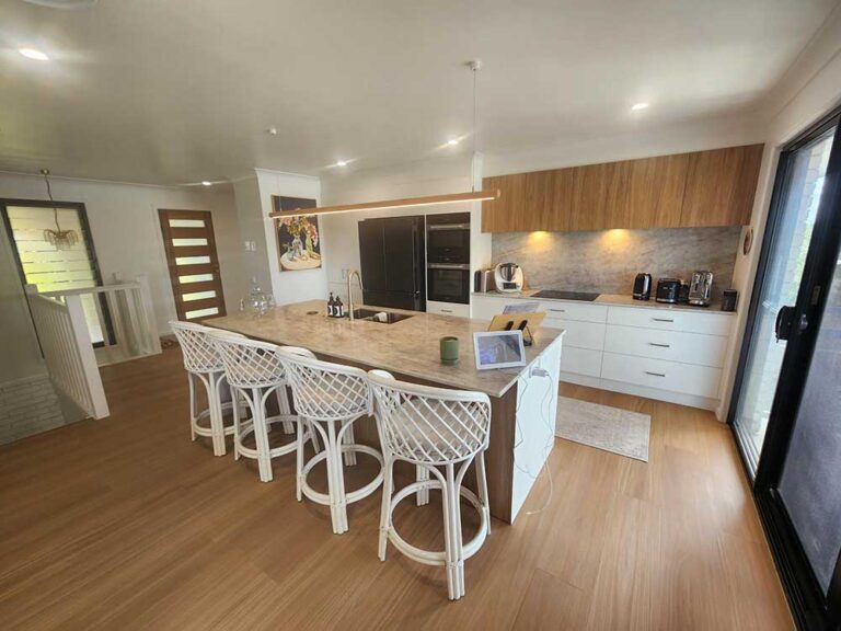 Bright, modern kitchen with a large marble island and six white wicker-bar stools, stainless steel appliances, and white lower cabinets beneath wood upper cabinets; sliding glass door to the outdoors on the right.