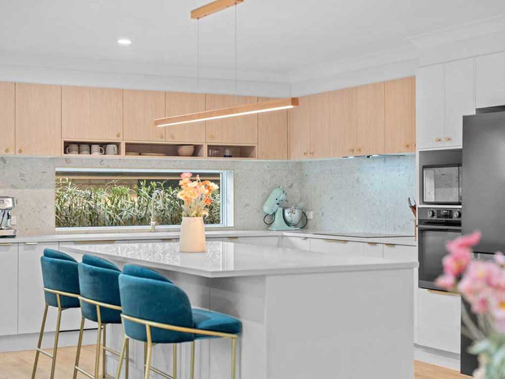 Modern kitchen with a white island, three teal bar stools, light wood upper cabinets, and a window overlooking greenery; a pastel teal mixer on the counter.