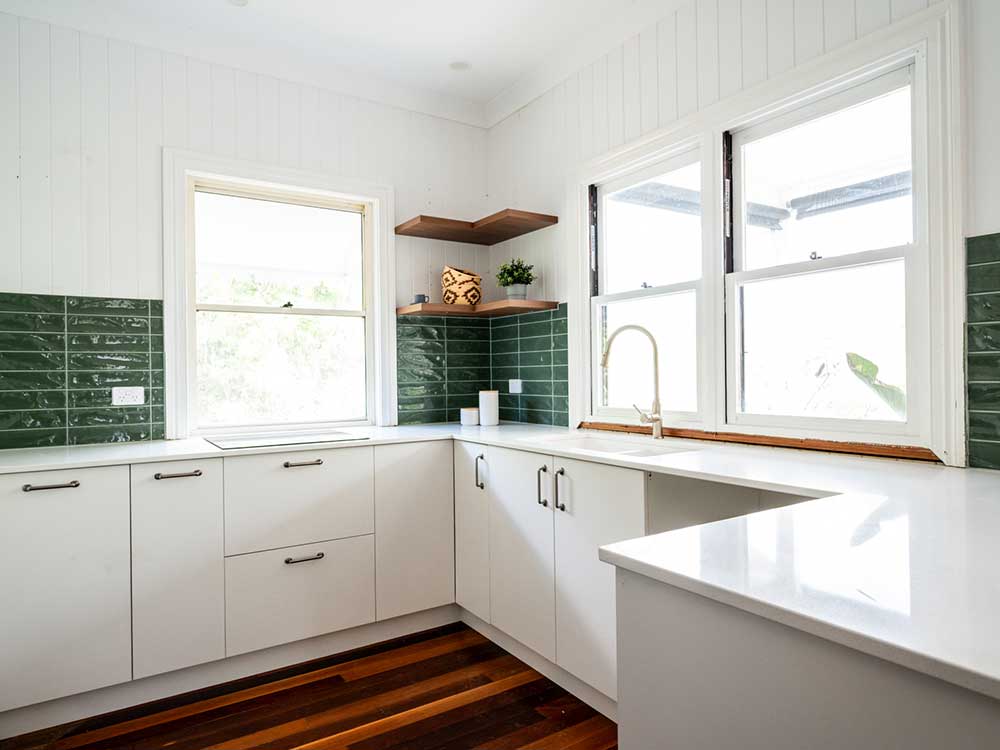 Bright white L-shaped kitchen with dark green subway tile backsplash, wooden shelves, and a stainless sink under large windows.